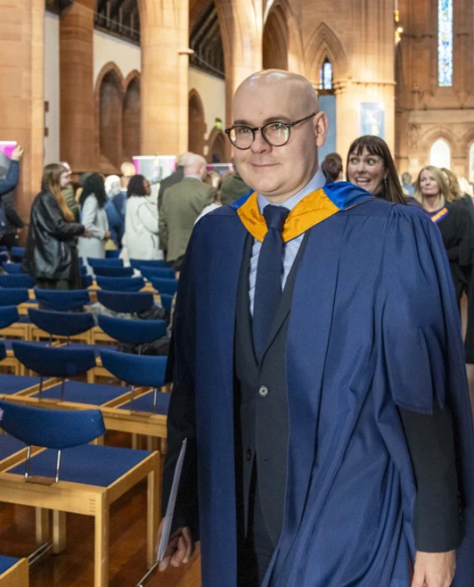 Lecturer in blue and gold academic gown smiling as they walk down the church aisle after the graduation ceremony. Lecturer in blue and gold academic gown smiling as they walk down the church aisle after the graduation ceremony.